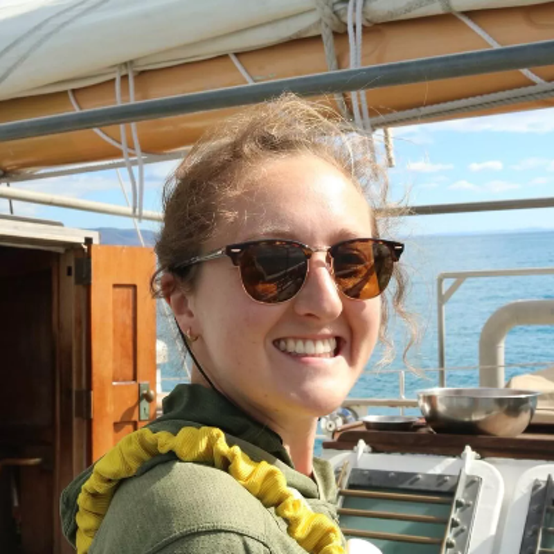 The image shows a young woman with sunglasses smiling on a boat. She is wearing a green jacket and a yellow life preserver. The background shows the ocean and the sky. She seems happy and relaxed. The boat appears to be a sailboat.